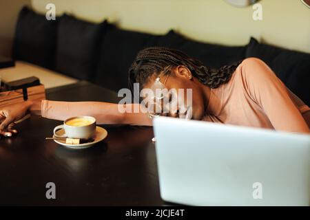 Ängstliche, unselbstbewusste multirassische afroamerikanische Frau in Brille, arbeiten online mit Laptop und Telefon. Kopfschmerzen, Migräne Stockfoto