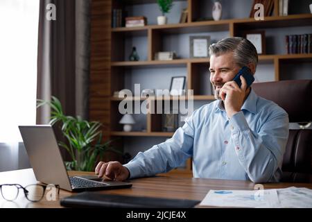 Porträt eines lächelnden Geschäftsmannes, der mit einem Laptop in der Büroeinrichtung telefoniert. Der männliche Händler verhandelt mit Geschäftspartnern und sucht nach neuen Stockfoto