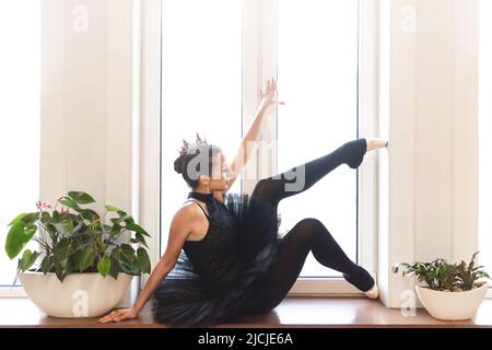 Anmutige Ballerina in schwarzem Schwanenkleid vor weißem Hintergrund. Junge Balletttänzerin, die vor der Aufführung in schwarzem Tutu, klassischem Tanzstud, praktiziert Stockfoto