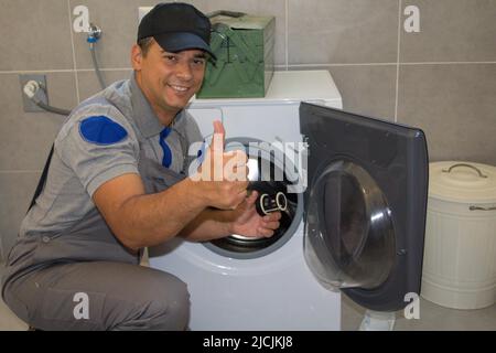 Foto eines lächelnden Klempners mit Daumen nach oben, als er eine defekte Waschmaschine repariert. Stockfoto