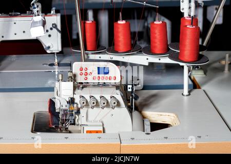 Industrielle Nähmaschine mit Spulen aus rotem Garn auf einem dunklen Hintergrund verschwommen Fabrik Nähwerkstatt. Stockfoto