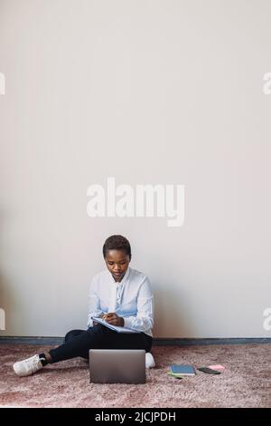 Afroamerikanische Geschäftsfrau, die während des Studiums Laptop benutzt und im Notebook schreibt und zu Hause auf dem Boden sitzt. Geschäftsplanung. Stockfoto