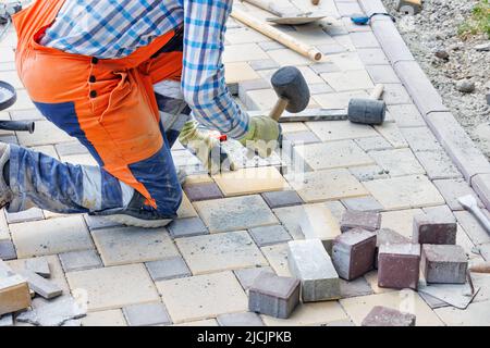 Ein Arbeiter in blauen und orangen Overalls hockt und legt Pflasterplatten mit einem Gummihammer. Nahaufnahme, selektiver Fokus, Kopierbereich. Stockfoto