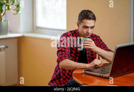 Ein junger Mann, der an einem Laptop arbeitet, sitzt neben einem Couchtisch mit einer Tasse heißen Tee. Arbeitsplatz in der Nähe des Fensters. Arbeiten Sie zu Hause. Stockfoto