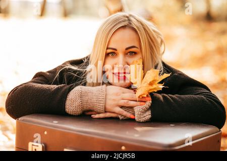 Herbstporträt der glücklichen, schönen 35 - jährigen blonden Frau in schwarzem Mantel und mit gelben Blättern in den Händen. Sie hält ihren Kopf in den Händen Stockfoto