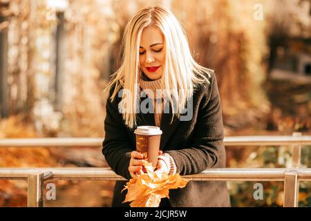 Strauß von hellen Blättern und Tasse Kaffee geben der schönen Frau in warmen, gemütlichen Kleidern am sonnigen Herbsttag im Stadtpark echte Freude. Freude teilen Stockfoto