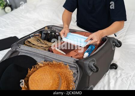 Mann Hand Verpackung Koffer Gepäck inklusive Gesichtsmaske und Alkoholspray zum Schutz Coronavirus (covid-19) auf einem Bett Stockfoto