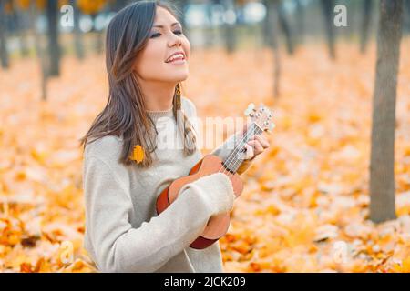 Junge schöne Mischrassenfrau, die Ukulele spielt und im Herbstpark singt Stockfoto