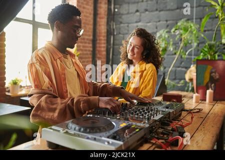 Fröhliche junge Frau, die mit Kopfhörern Musik hört und ihren Freund vor dem dj-Set sieht und neue Musik kreiert Stockfoto