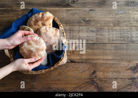 Die Hände der Frau legen hausgemachtes Pita-Brot in einen Holzkorb auf einen alten Holztisch. Speicherplatz kopieren. Stockfoto