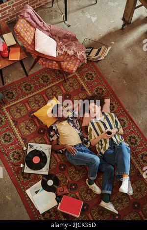 Junge Frau mit einer Tasse schwarzen Tee oder Kaffee, die mit ihrem Freund spricht, während sie sich beide auf dem roten Teppich ausruhen und Vynil-Scheiben lauschen Stockfoto