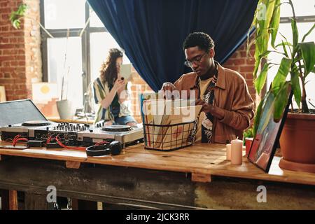 Der junge afroamerikanische Mann schaut sich seine Sammlung von Vynil-Disks in einer Kiste an, die auf dem Tisch neben der Deejay-Kabine und den Kopfhörern steht Stockfoto