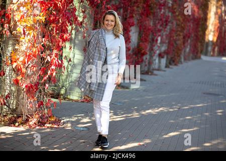 Charmante Blondine in leichter Hose, Kaschmirpullover und graue Tweed-Jacke drapiert über ihre Schulter geht zu Fuß durch herbstliche Straßen der Stadt. Hell Stockfoto