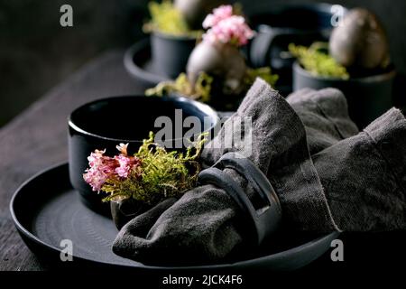 Ostertisch mit leeren schwarzen Keramikplatten und -Tassen, verziert mit Waldmoos, schwarzen Ostereiern und rosa Frühlingsblumen auf Holz-T Stockfoto