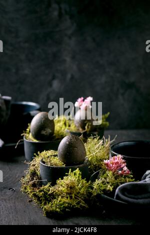 Ostertisch mit leeren schwarzen Keramikplatten und -Tassen, verziert mit Waldmoos, schwarzen Ostereiern und rosa Frühlingsblumen auf Holz-T Stockfoto