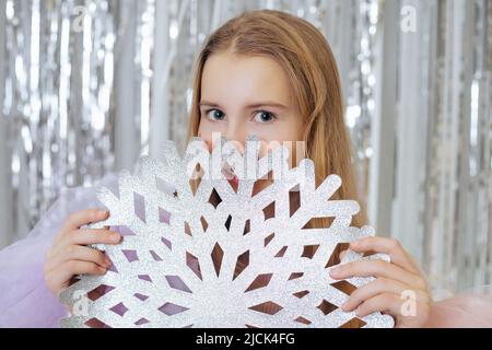 Das schöne Teenager-Mädchen lächelt geheimnisvoll und versteckt sich hinter einer großen Schneeflocke aus Papier, die sie in ihren Händen hält. Neujahr und Weihnachten. Dekoration und Stockfoto