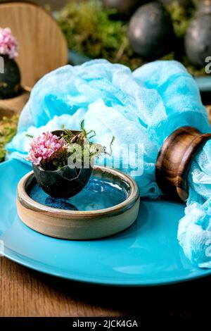 Ostertisch mit leeren, türkisfarbenen Keramikplatten, verziert mit Waldmoos, schwarzen Ostereiern und rosa Frühlingsblumen auf einem Holztisch. Stockfoto