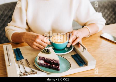 Die Hände reifer Frauen halten eine Tasse Cappuccino und einen Sahnetorte. Erwachsene Dame in weißem Pullover genießen ihr Frühstück Latte und Dessert Pie auf Stockfoto