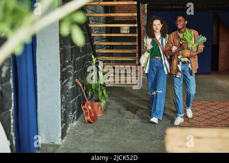 Glückliches junges interkulturelles Paar in stilvoller Casualwear, das sich im Wohnzimmer der Loft-Wohnung bewegt, während es vom Supermarkt zurückkommt Stockfoto