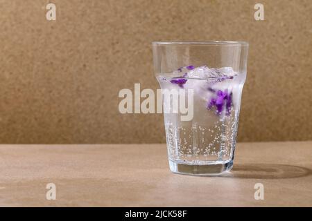 Transparentes Glas Limonade mit Veilchen-Blumeneiswürfeln für kalte sommerliche Erfrischung, Limonadencocktail. Auf brauner Lasche stehend Stockfoto