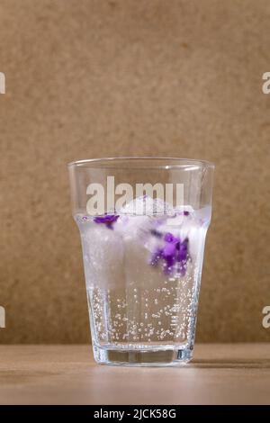 Transparentes Glas Limonade mit Veilchen-Blumeneiswürfeln für kalte sommerliche Erfrischung, Limonadencocktail. Auf brauner Lasche stehend Stockfoto