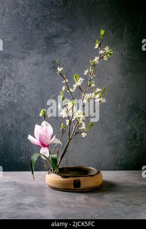 Frühling Ikebana. Florale Komposition mit frühlingsblühenden Magnolien und Pflaumenzweigblumen in brauner Keramikschale, die auf einem grauen Tisch steht. Japanischer Stil Stockfoto