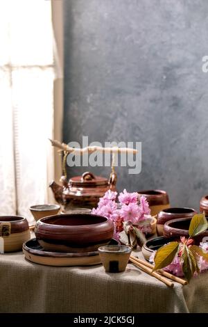 Japanische Tischdekoration im asiatischen Stil mit leerem Keramikgeschirr, braunen, rauen Schüsseln, Wasserkocher und Tassen auf Leinentischdecke, dekoriert mit rosa Sprossen Stockfoto