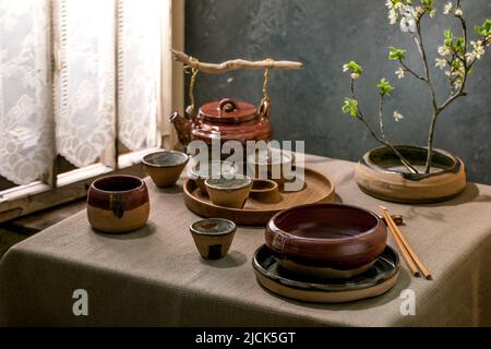 Japanische Tischdekoration im asiatischen Stil mit leerem Keramikgeschirr, braunen, rauen Schalen und Tassen auf Leinentischdecken, dekoriert von ikebana Spring blo Stockfoto