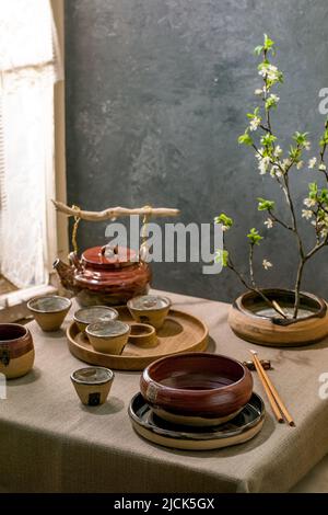 Japanische Tischdekoration im asiatischen Stil mit leerem Keramikgeschirr, braunen, rauen Schalen und Tassen auf Leinentischdecken, dekoriert von ikebana Spring blo Stockfoto