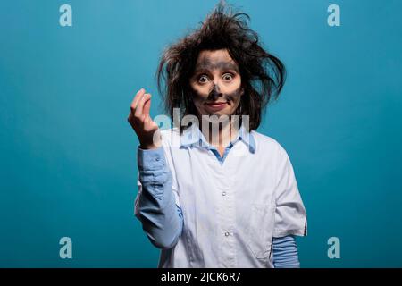 Dumm aussehende verrückte Chemikerin mit schmutzigem Gesicht und unordentlichen Haaren, die auf blauem Hintergrund auf die Kamera schaut. Verrückter Wissenschaftler mit urkomischer, witziger Art, sich um die Explosion von Laborchemikalien zu kümmern. Studioaufnahme. Stockfoto