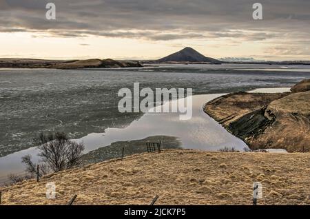 Blick während des Sonnenuntergangs über den teilweise gefrorenen See Myvatn in Island, umgeben von grasbewachsenen Hügeln und Mount Vindbelgjarfall Stockfoto