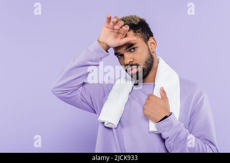 Müde afroamerikanische Mann in Pullover mit weißem Handtuch und wischen Schweiß mit Hand isoliert auf lila Stockfoto