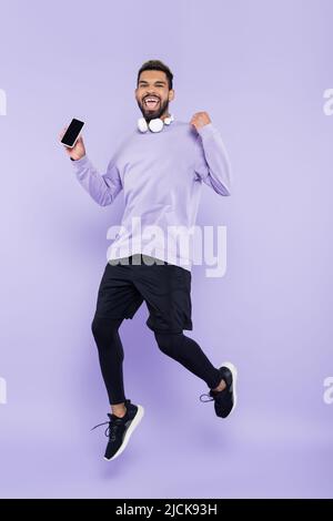 Aufgeregter afroamerikanischer Mann mit kabellosen Kopfhörern, die das Smartphone halten und auf lila springen Stockfoto