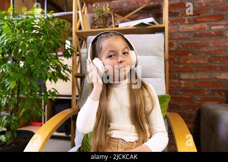 Portrait von schönen kleinen Mädchen, Schüler im Schulalter zu Hause studieren, Buch lesen. Modell mit Kopfhörern. Bildung, Kindheit, Menschen, Hausaufgaben und Stockfoto
