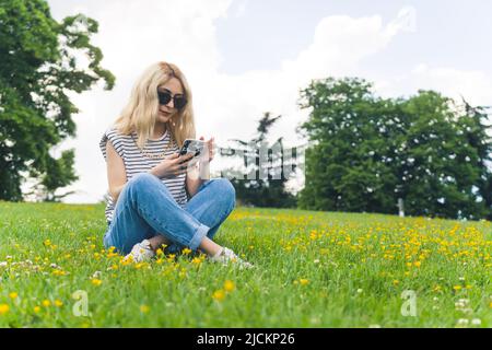 Hübsches kaukasisches Mädchen in legerer Kleidung und Sonnenbrille, das auf dem Gras sitzt und ein Telefon benutzt, voller Schuss im Park. Hochwertige Fotos Stockfoto