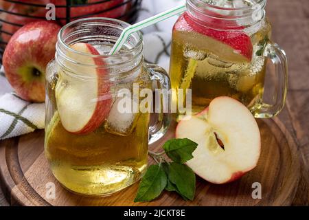 Zwei Gläser Apfelsaft mit Apfelscheiben und Eis auf einer Holzplatte mit einem Korb mit Äpfeln im Hintergrund Stockfoto