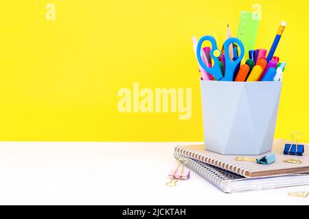 Bildungskonzept - Notizbücher, Schreibwaren und grauer Organizer mit mehrfarbigen Stiften, Markern, Bleistiften und Scheren auf einem weißen Tisch im Klassenzimmer. Stockfoto