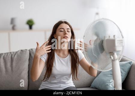 Die traurige junge europäerin, die unter Hitze leidet, sitzt auf dem Sofa, fängt kalte Luft vom Ventilator und winkt die Hände zu sich selbst. Stockfoto