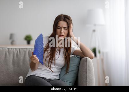 Traurige junge europäische Frau leidet unter heißen und Kopfschmerzen bläst Fan auf sich selbst sitzen auf dem Sofa im Wohnzimmer Stockfoto