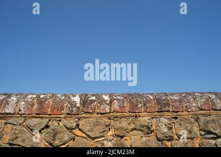 Kentish Ragstone ein harter grauer Kalkstein, der verwendet wurde, um eine starke Grenzmauer mit roten Ziegelsteinen zu errichten, die oben gegen einen blauen Himmel stehen Stockfoto