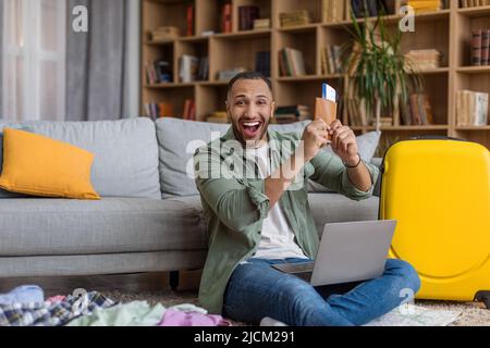 Überglücklich afroamerikanischer Mann, der Pass und Flugticket vorzeigte und mit einem Laptop Touristenurlaub kaufte Stockfoto