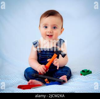 6 Monate alter Junge, der in Denim-Overalls gekleidet ist, während er als Werkzeugarbeiter spielt. Er sieht aus wie ein kleiner Zimmermann oder ein kleiner Klempner. Stockfoto