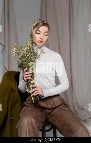 Stilvolle Frau im weißen Rollkragen sitzend mit Blumenstrauß aus Gypsophila auf grauem Hintergrund Stockfoto