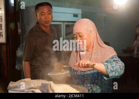 Frauen mittleren Alters, die muslimische Kopfbedeckung tragen, servieren in einer lokalen Restaurantküche heißes, dampfendes chinesisches Essen zu einem Gericht, das einem Kunden serviert wird und zu essen gibt. XI'an, China. (125) Stockfoto