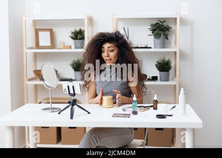 Fröhlich gebräunt schöner lockiger lateinischer Beauty-Blogger in Casual Top erzählt von Körpercreme im Heim-Interieur. Space Mockup Banner kopieren. Influencer-Rekord Stockfoto