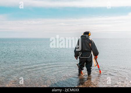 Ein Mann, der knietief im Wasser steht und mit einem Metalldetektor nach Edelmetallen sucht. Meer und Himmel im Hintergrund. Stockfoto