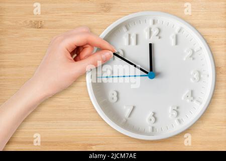 Die Hand einer Frau bewegt die Zeiger der Uhr aus nächster Nähe. Flach liegend. Holzhintergrund. Das Konzept des Zeitmanagements. Stockfoto