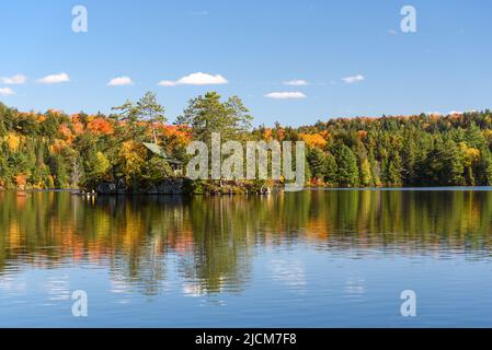 Hütte auf einer kleinen felsigen Insel in der Mitte eines Sees, umgeben von dichtem Wald an einem sonnigen Herbsttag. In der Nähe der Insel befinden sich Kanufahrer. Stockfoto