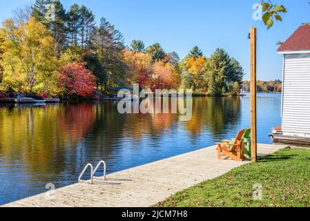 Verlassene Holzpromenade am Flussufer mit zwei Adirondack-Stühlen an einem sonnigen Herbsttag. Herbstfarben spiegeln sich im Wasser. Stockfoto