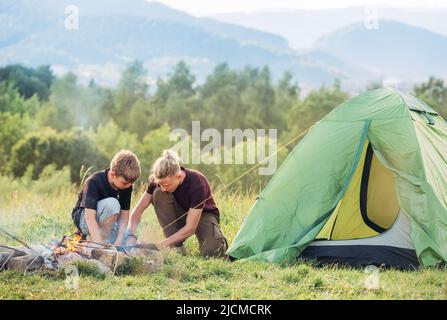 Zwei Brüder im Teenageralter, die das grüne Zelt neben dem Lagerfeuer aufschlagen. Aktive Zeit im Freien verbringen oder Camping in der Natur Konzept. Stockfoto